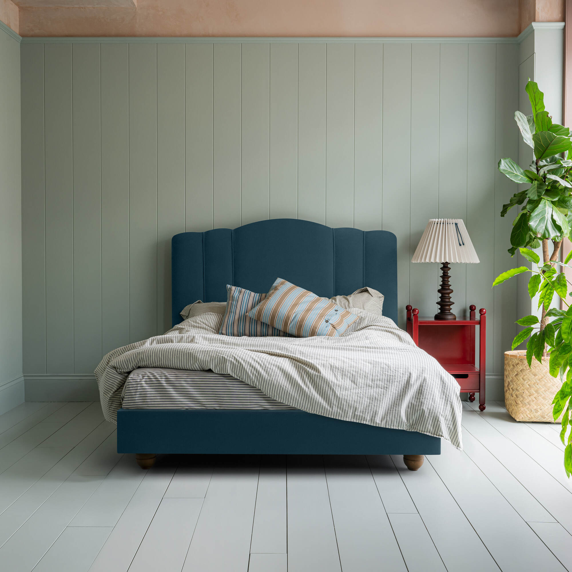 Cloud Nine upholstered bed in Intelligent Velvet Aegean, styled without a valance, featuring a red side table, lamp, blanket, and cushions, designed by Nicola Harding & Co.