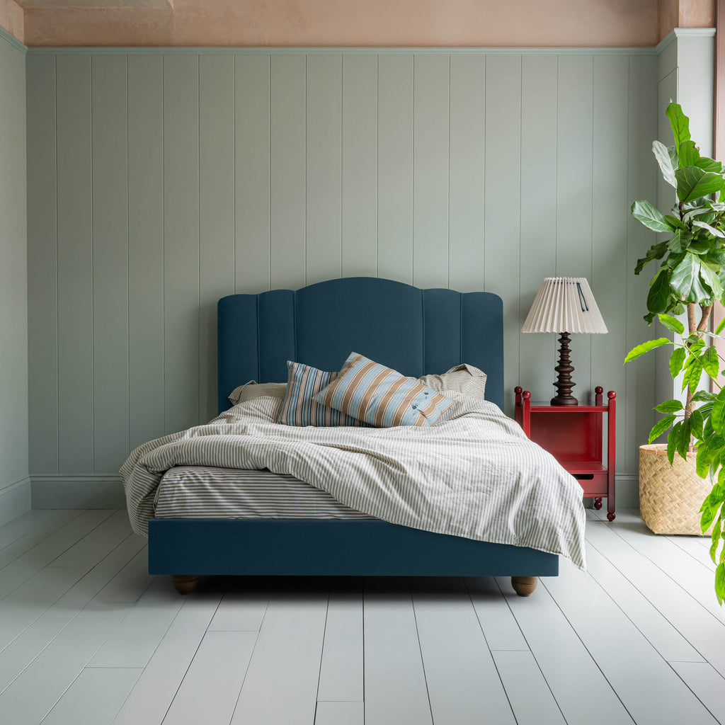 Cloud Nine upholstered bed in Intelligent Velvet Aegean, styled without a valance, featuring a red side table, lamp, blanket, and cushions, designed by Nicola Harding & Co.
