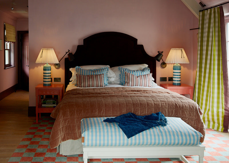 A bedroom featuring a stunning black velvet bed paired with a vibrant red side table and a blue base lamp emitting warm, ambient light, creating a cozy and elegant atmosphere. Designed by Nicola Harding.