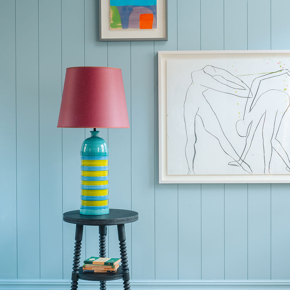 Humbug Straight Empire Paper Lampshade in Burgundy Paper with Pink Trim, With Humbug Striped Ceramic Table Lamp Base in Blue & Warm Yellow, On Spindle Side Table, Carbon Black, With Beautiful Art Hanging on the Wall in Back-Ground- Nicola Harding