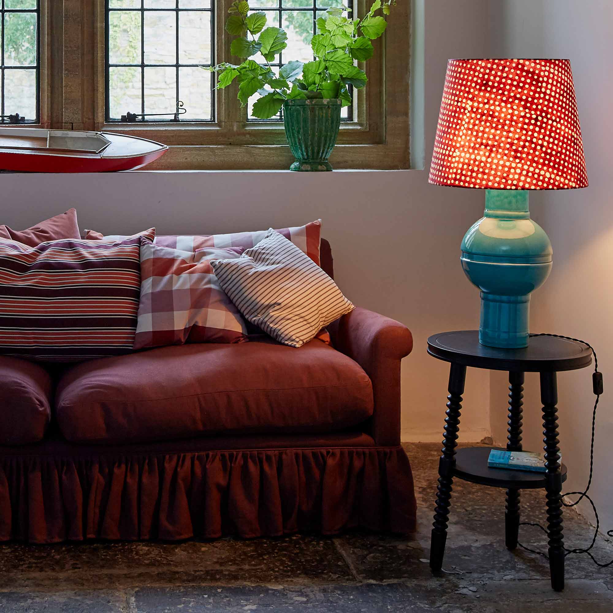 Humbug Straight Empire Batik Paper Lampshade in Dotty Maroon with Hourglass Ceramic Table Lamp Base in Blue, Lights 'ON,' Next to NiX 4-Seater Sofa and Spindle Side Table in Carbon Black – Nicola Harding