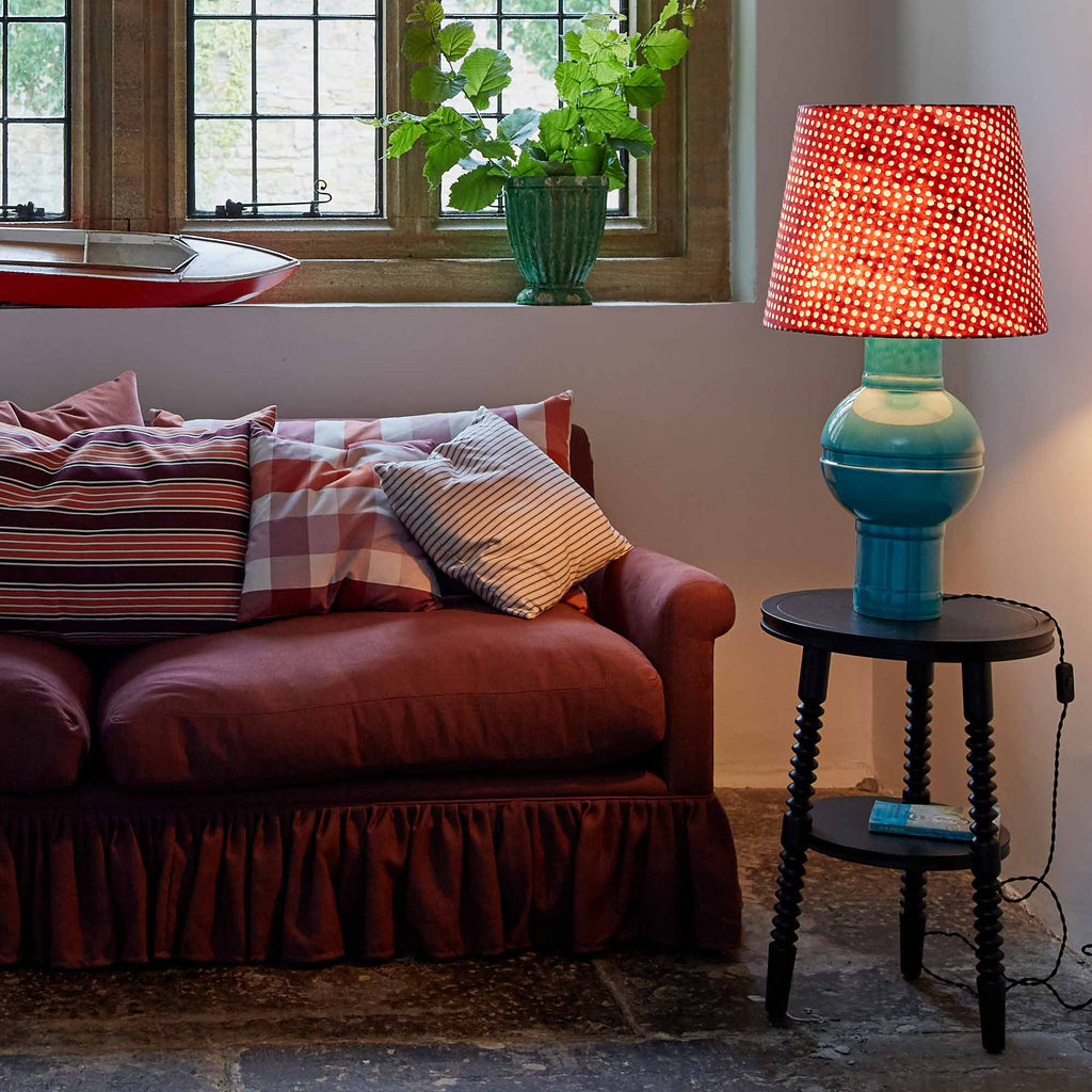 Humbug Straight Empire Batik Paper Lampshade in Dotty Maroon with Hourglass Ceramic Table Lamp Base in Blue, Lights 'ON,' Next to NiX 4-Seater Sofa and Spindle Side Table in Carbon Black – Nicola Harding