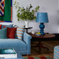 Humbug Straight Empire Batik Paper Lamp Shade in Dotty Sky Blue, With Hourglass Ceramic Table Lamp Base in Blue, On a Side Table Next to Ottomans and NiX Velvet Sofa With Cushions - Nicola Harding
