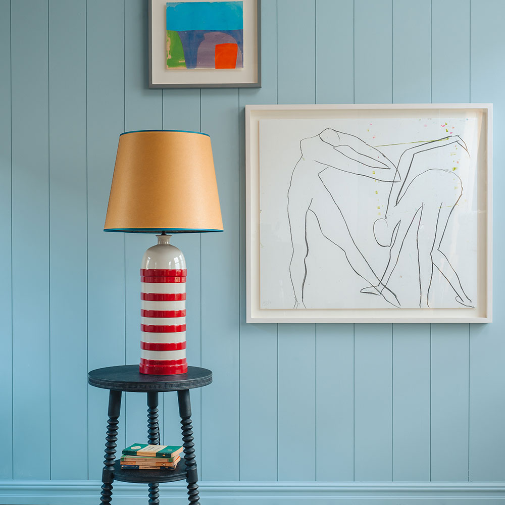 Humbug Striped Ceramic Table Lamp Base in Cherry Red & Warm White, With Humbug Straight Empire Paper Lamp Shade in Mustard with Blue Trim, On a Spindle Side Table, Carbon Black, With Beautiful Art Hanging on the Wall - NiX By Nicola Harding & Co.