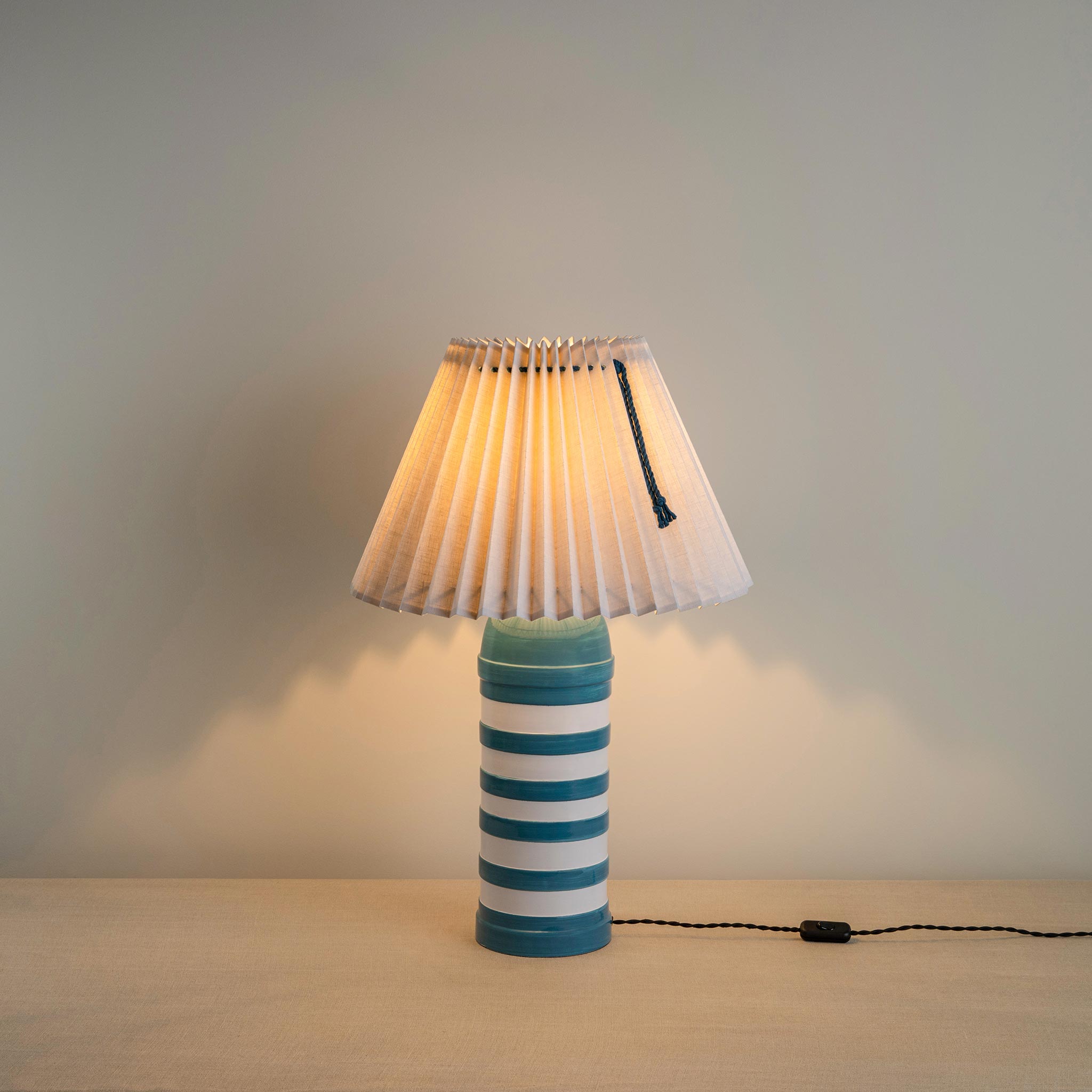 Humbug Striped Ceramic Table Lamp Base in Blue & Warm White With Sunburst Concertina Natural Pleat Lamp Shade with Peacock Blackberry Cord, With Lights 'ON' - Nicola Harding