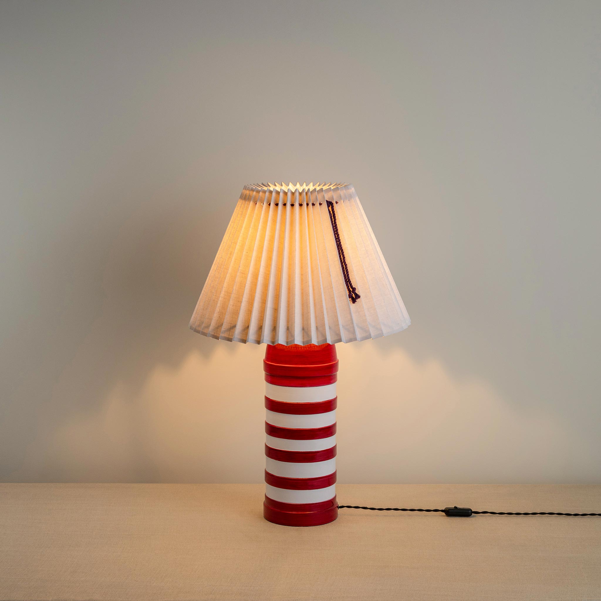 Humbug Striped Ceramic Table Lamp Base in Cherry Red & Warm White, With Sunburst Concertina Natural Pleat Lamp Shade with Blackberry Cord, With Lights 'ON' - Nicola Harding