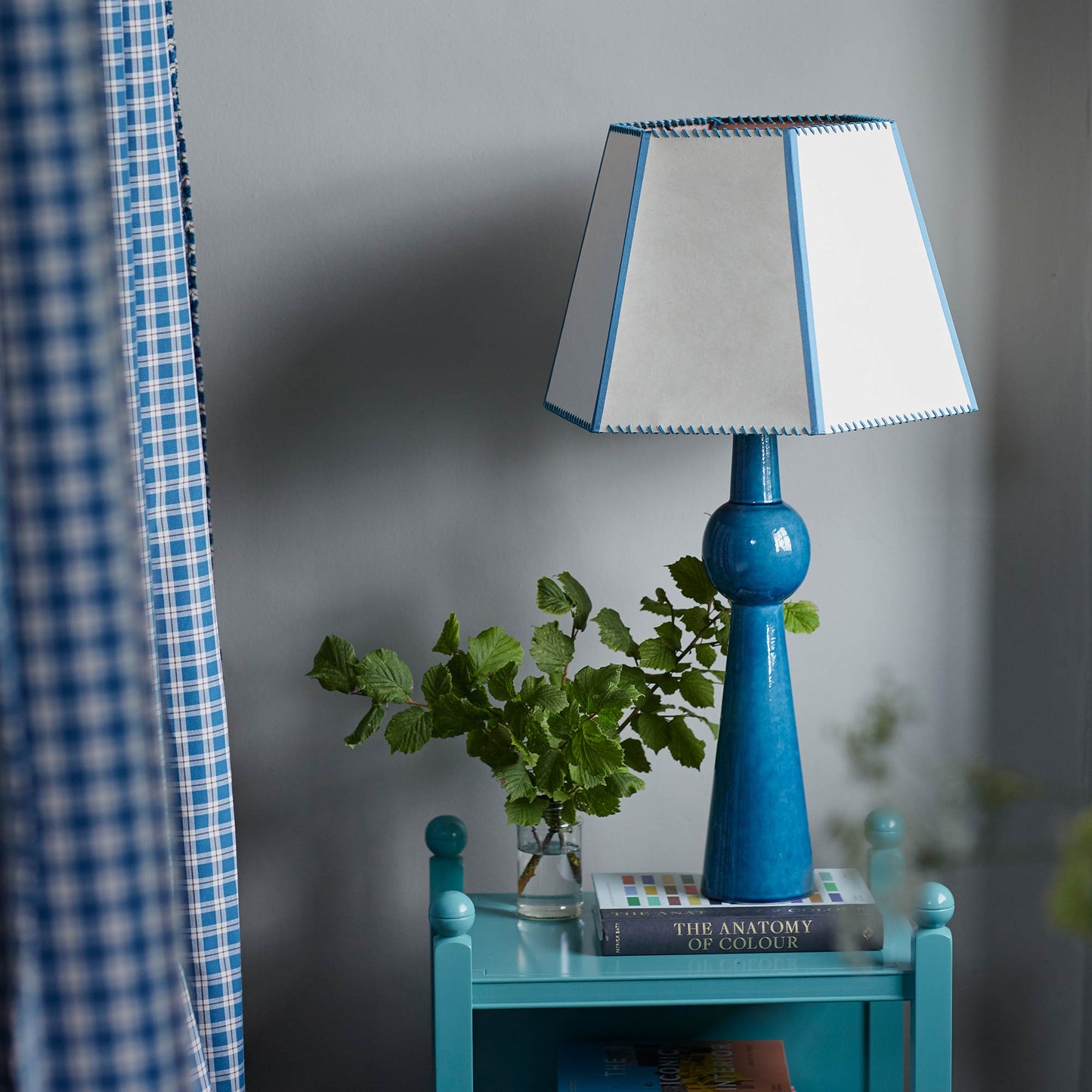 Comet Ceramic Table Lamp Base in Blue, With Townhouse Hexagonal Lamp Shade in Soft White with Peacock Blue Trim & Stitching, On a Blue Sidetable With a Beautiful Plant - Nicola Harding