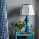 Comet Ceramic Table Lamp Base in Blue, With Townhouse Hexagonal Lamp Shade in Soft White with Peacock Blue Trim & Stitching, On a Blue Sidetable With a Beautiful Plant - Nicola Harding