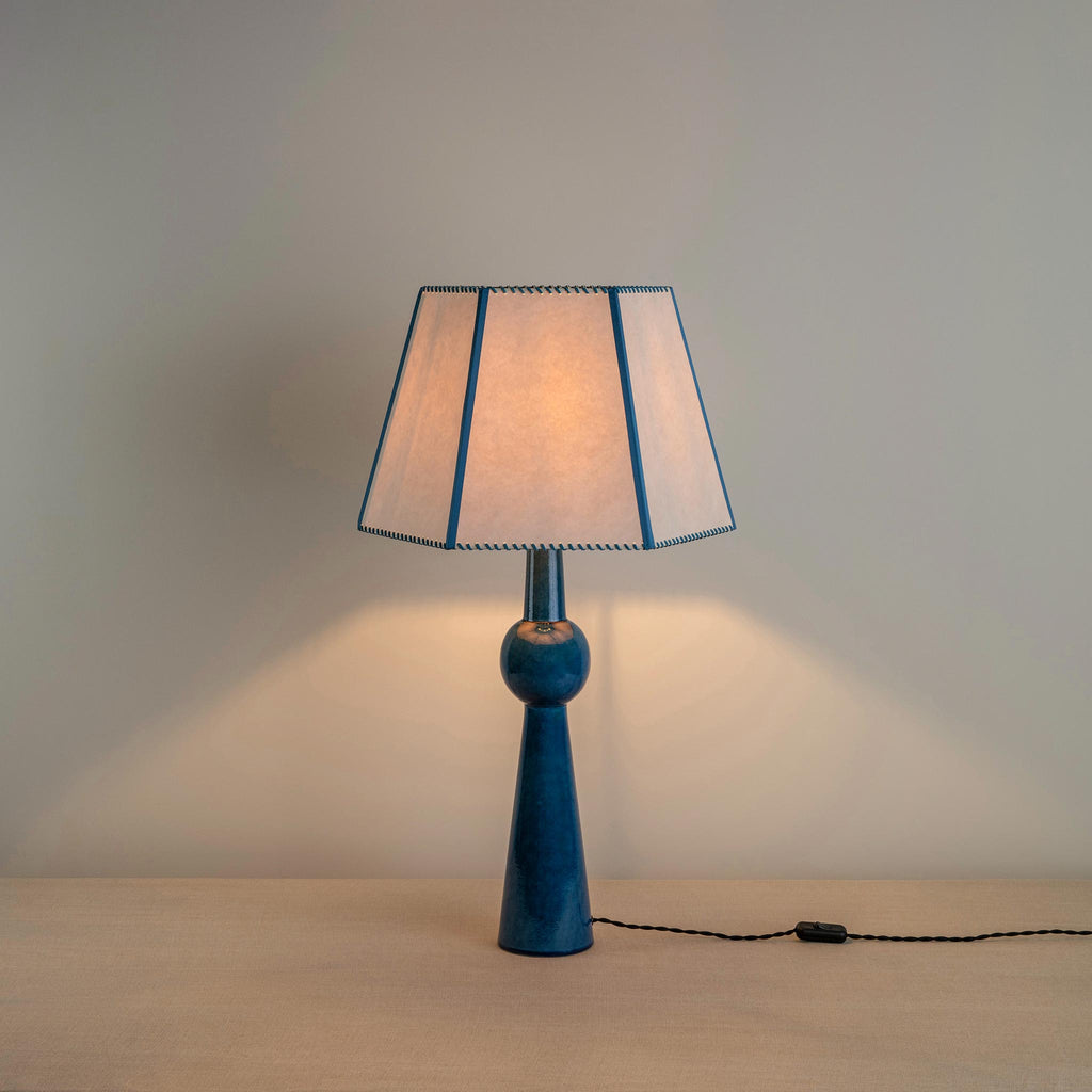 Comet Ceramic Table Lamp Base in Blue, With Townhouse Hexagonal Lamp Shade in Soft White with Peacock Blue Trim & Stitching, With Lights 'ON' - Nicola Harding