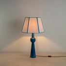 Comet Ceramic Table Lamp Base in Blue, With Townhouse Hexagonal Lamp Shade in Soft White with Peacock Blue Trim & Stitching, With Lights 'ON' - Nicola Harding