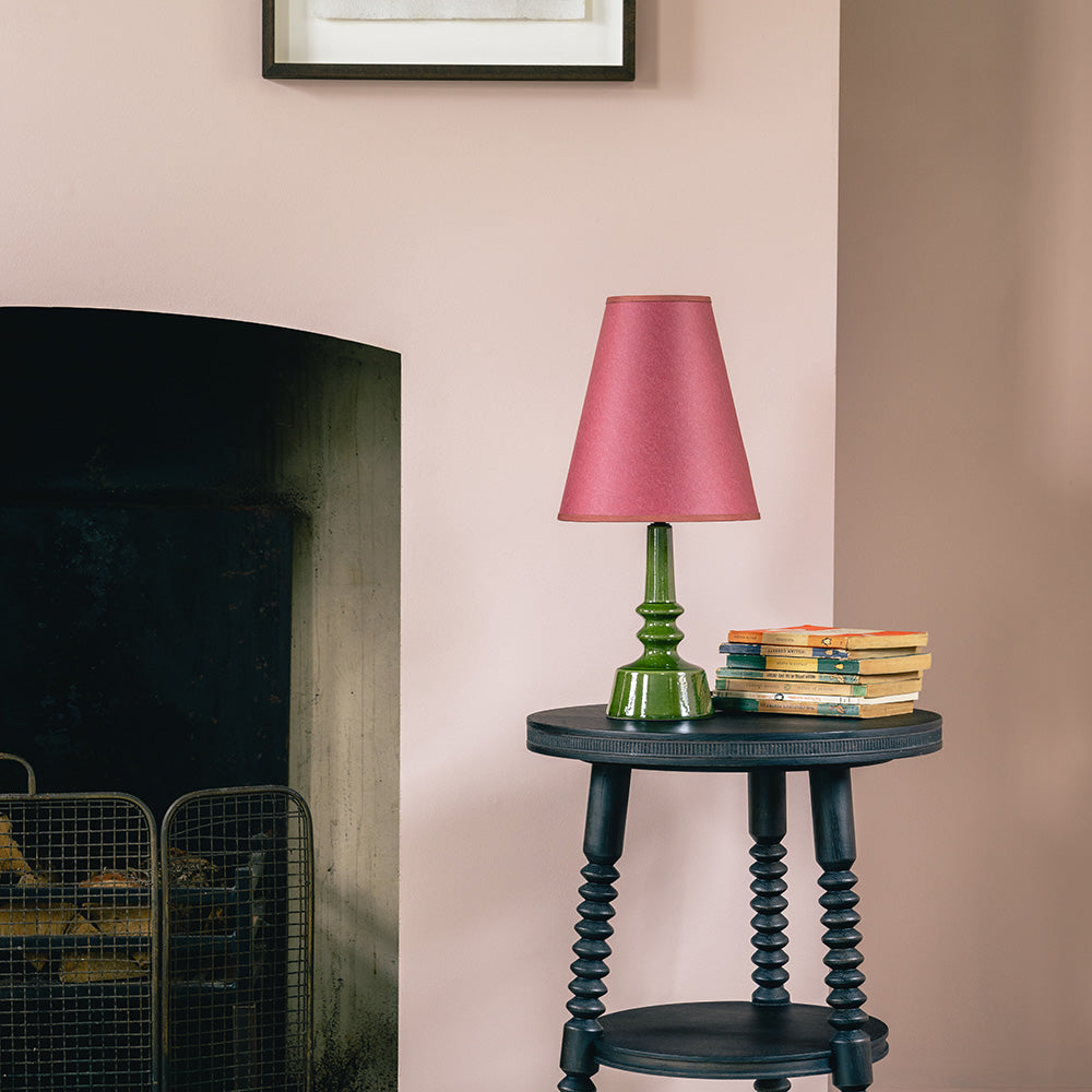 Ditsy Ceramic Table Lamp Base in Olive Green, With Focused Empire Paper Lamp Shade in Burgundy with Muted Pink Trim On a Black Side Table Near a Fire Place With a Couple of Books - Nicola Harding