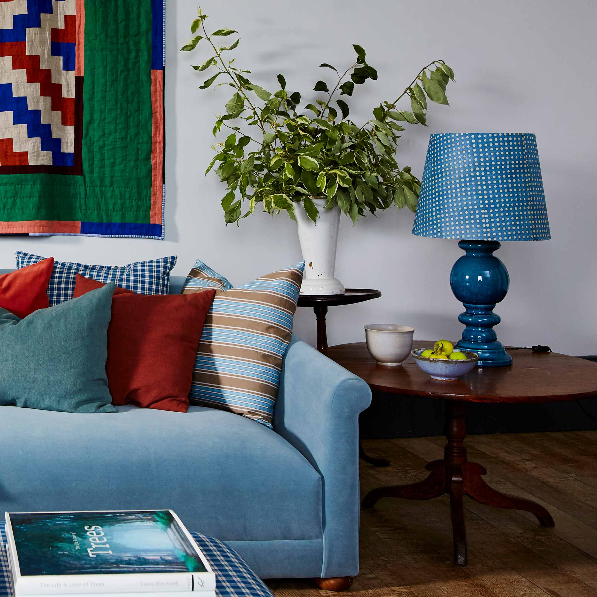 Hourglass Ceramic Table Lamp Base in Blue, With Humbug Straight Empire Batik Paper Lamp Shade in Dotty Sky Blue, On a Side Table Along With The NiX Velvet Sofa With Multiple Cushions on-it - Nicola Harding