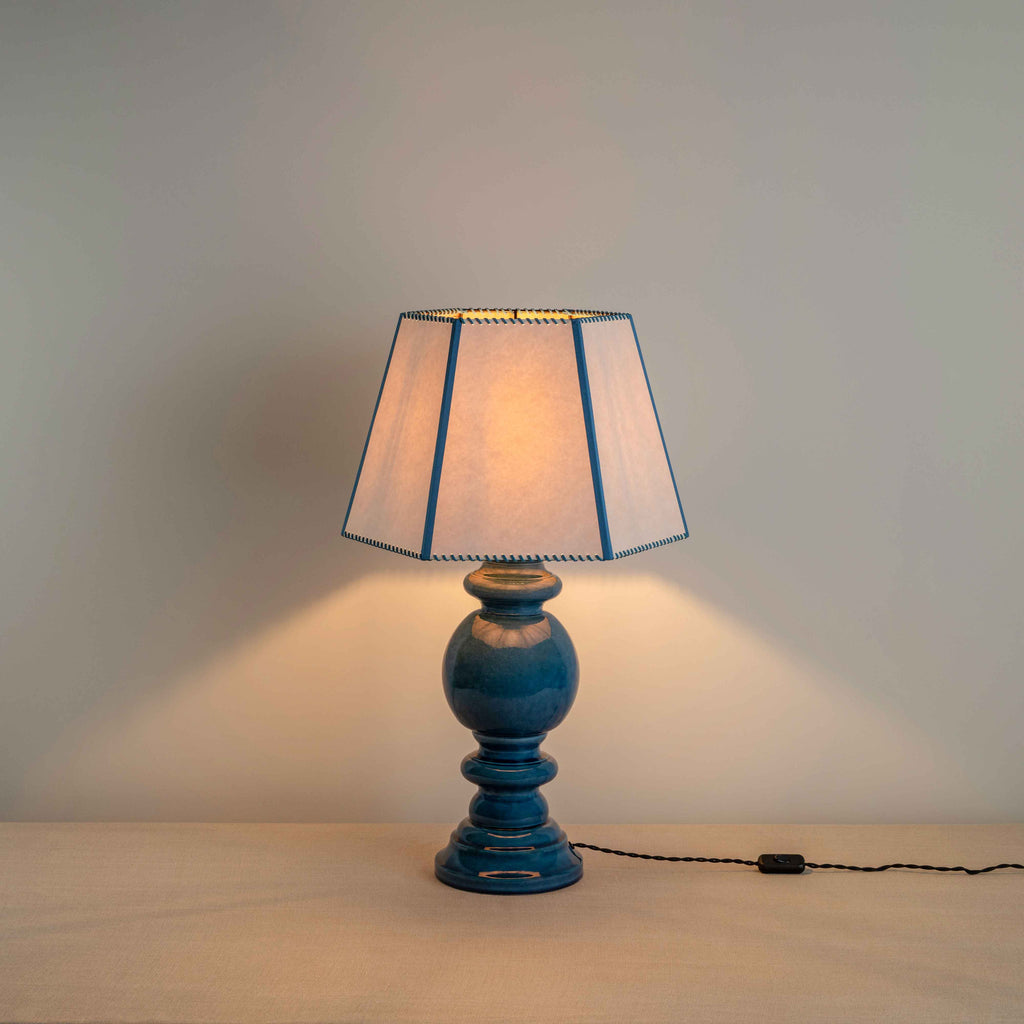 Hourglass Ceramic Table Lamp Base in Blue, With Townhouse Hexagonal Lamp Shade in Soft White with Peacock Blue Trim & Stitching, With Lights 'ON' - Nicola Harding