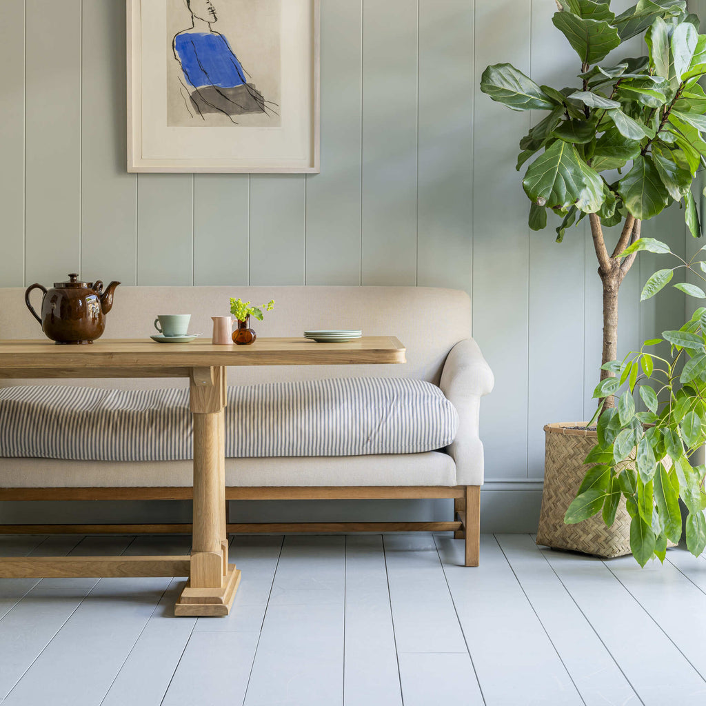 Front Row upholstered bench in Laidback Linen Dove frame, with a dining table in front featuring a teapot, cup, and a bunch of plates, designed by Nicola Harding.