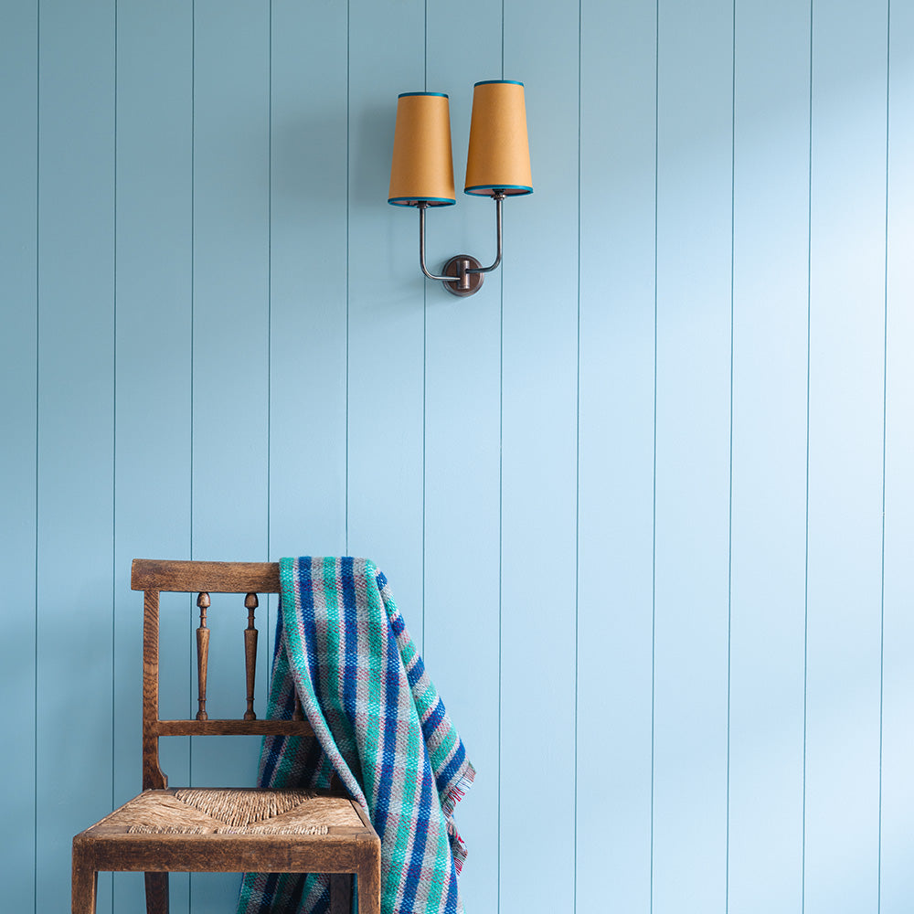 Double Trouble Waxed Brass Wall Light With Mustard Lamp Shades, With Light Blue Wall and a Chair With a Wool Blanket On-it - Nicola Harding