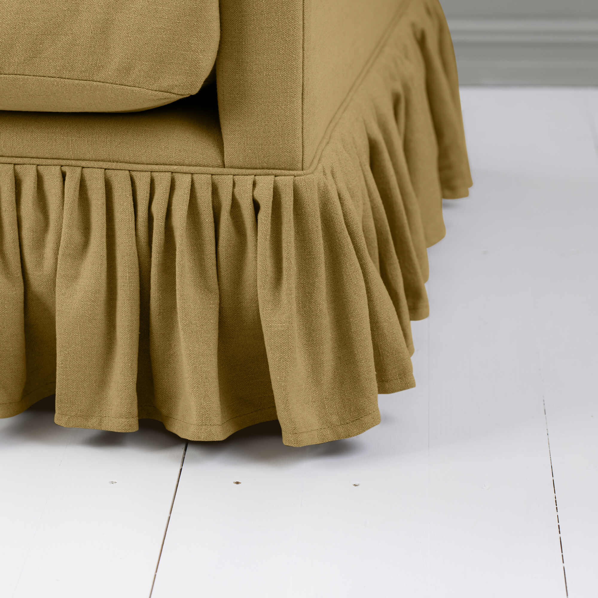  Close-up of the bottom corner of a beige upholstered sofa with a ruffled skirt. 