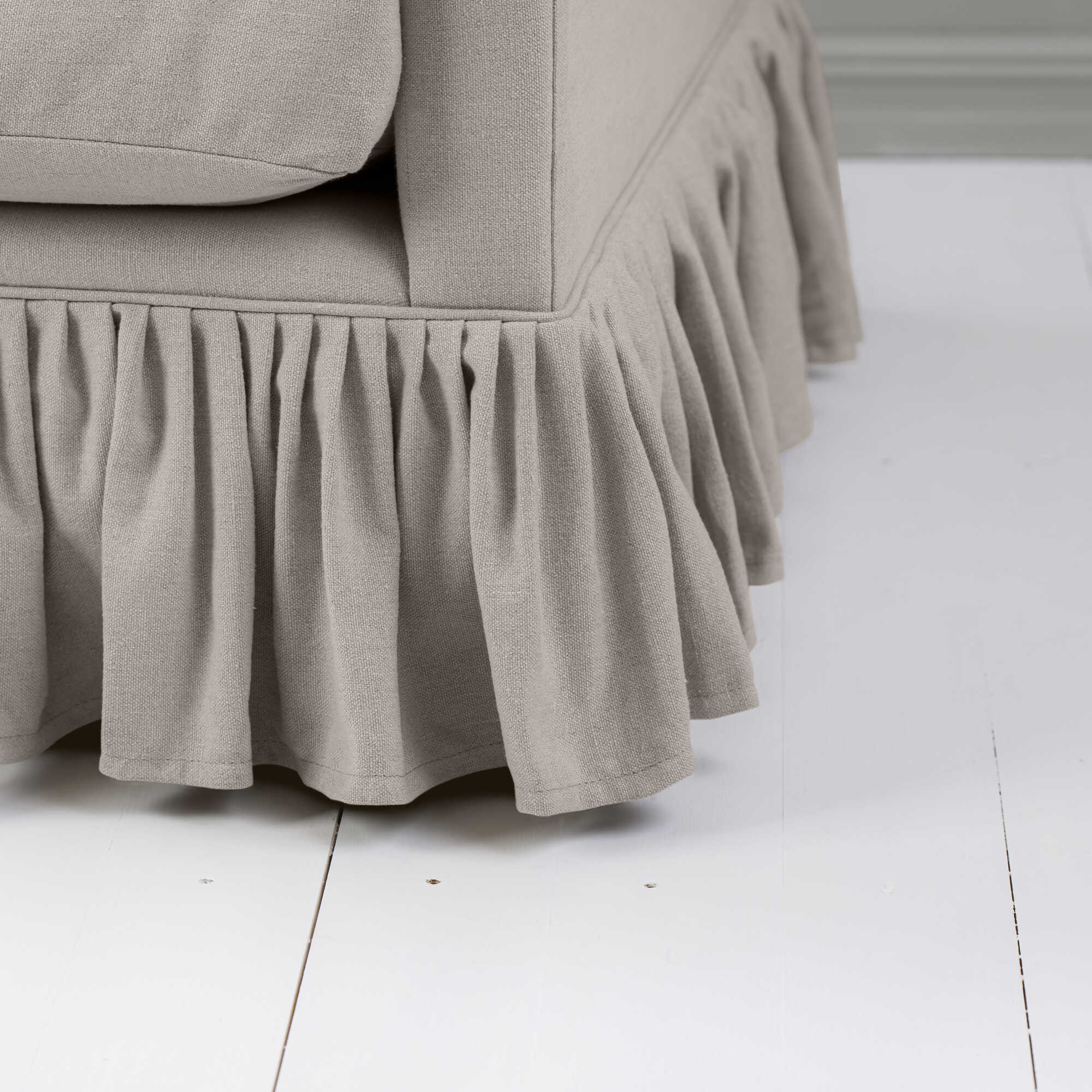  Close-up of a gray fabric sofa with a pleated skirt detail, set on a white wooden floor. 