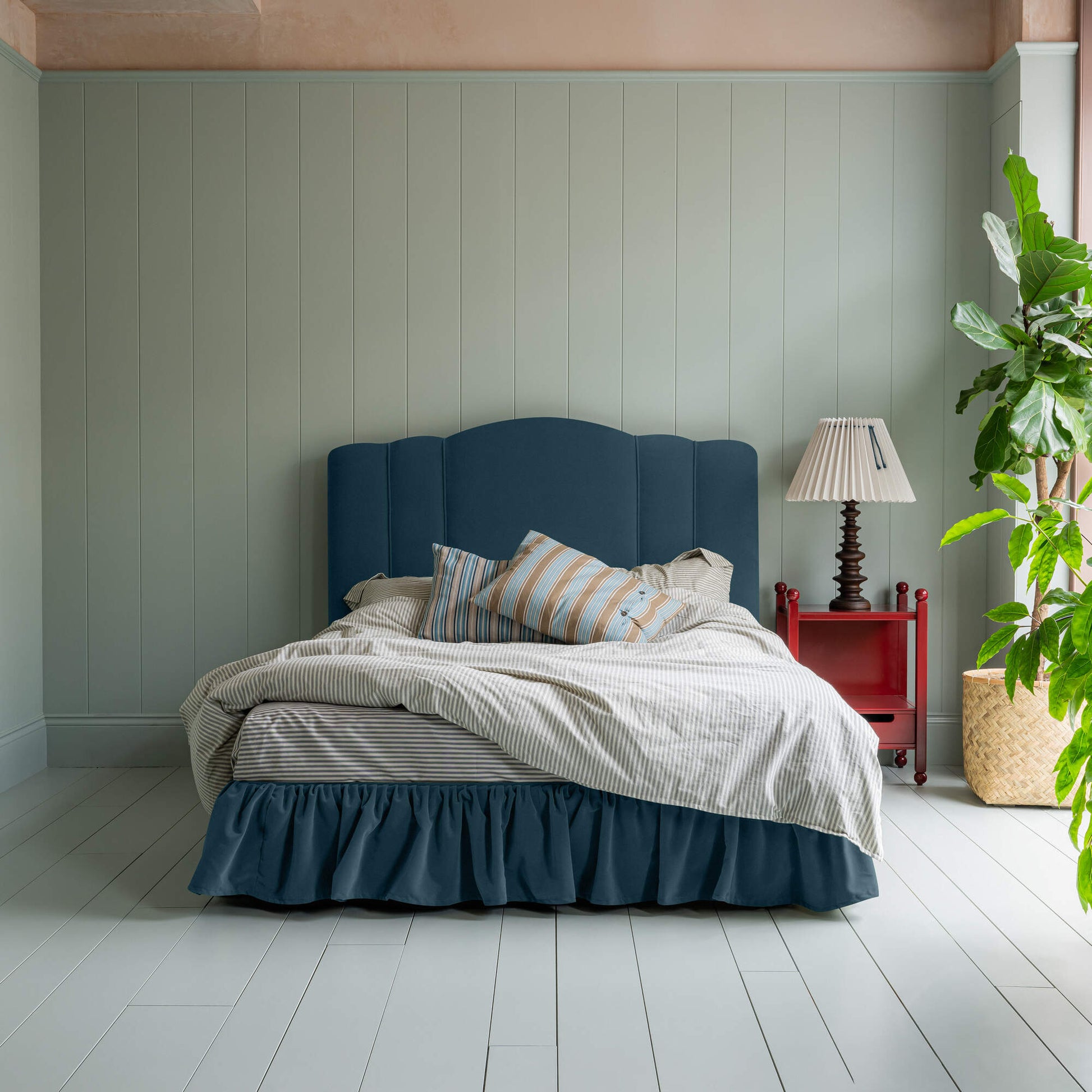 Cloud Nine Upholstered Bed in Intelligent Velvet Aegean with Ruffled Valance, accompanied by a red side table and a  lamp, pillows, and a blanket.