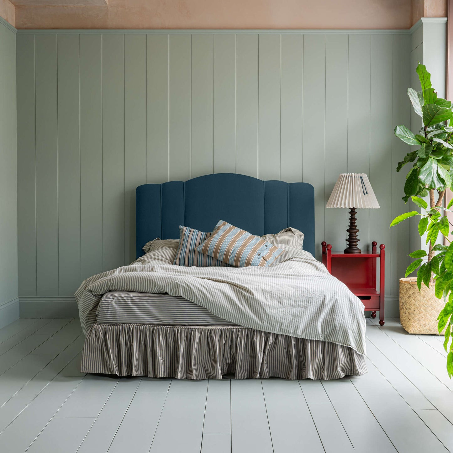 Cloud Nine Upholstered Bed in Intelligent Velvet Aegean with Ticking Cotton Ruffled Valance, accompanied by a red side table with a lamp, pillows, and a blanket.