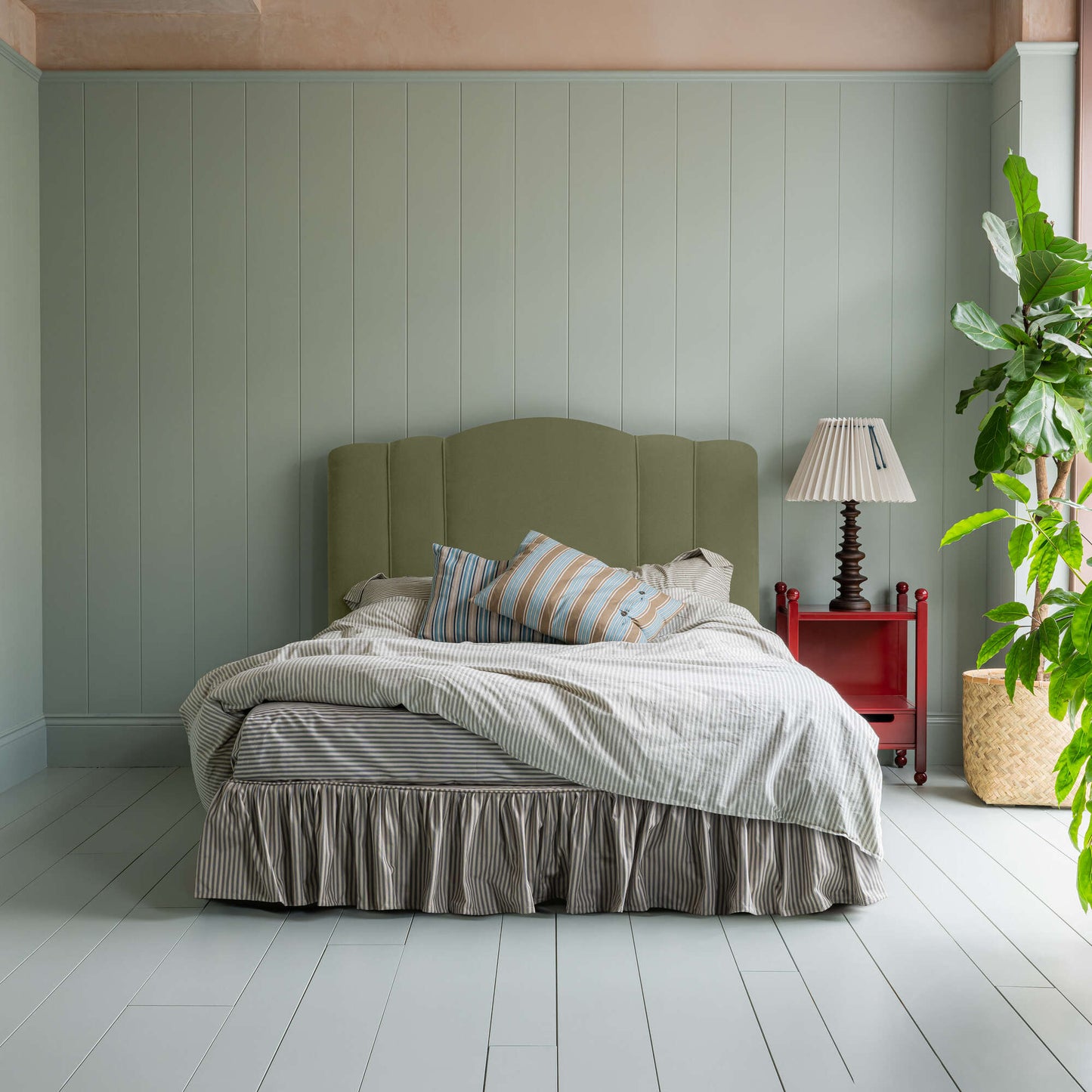 Bed in Intelligent Velvet Green Tea with Ticking Cotton Ruffled Valance, accompanied by a red side table and a lamp, pillows, and a blanket.