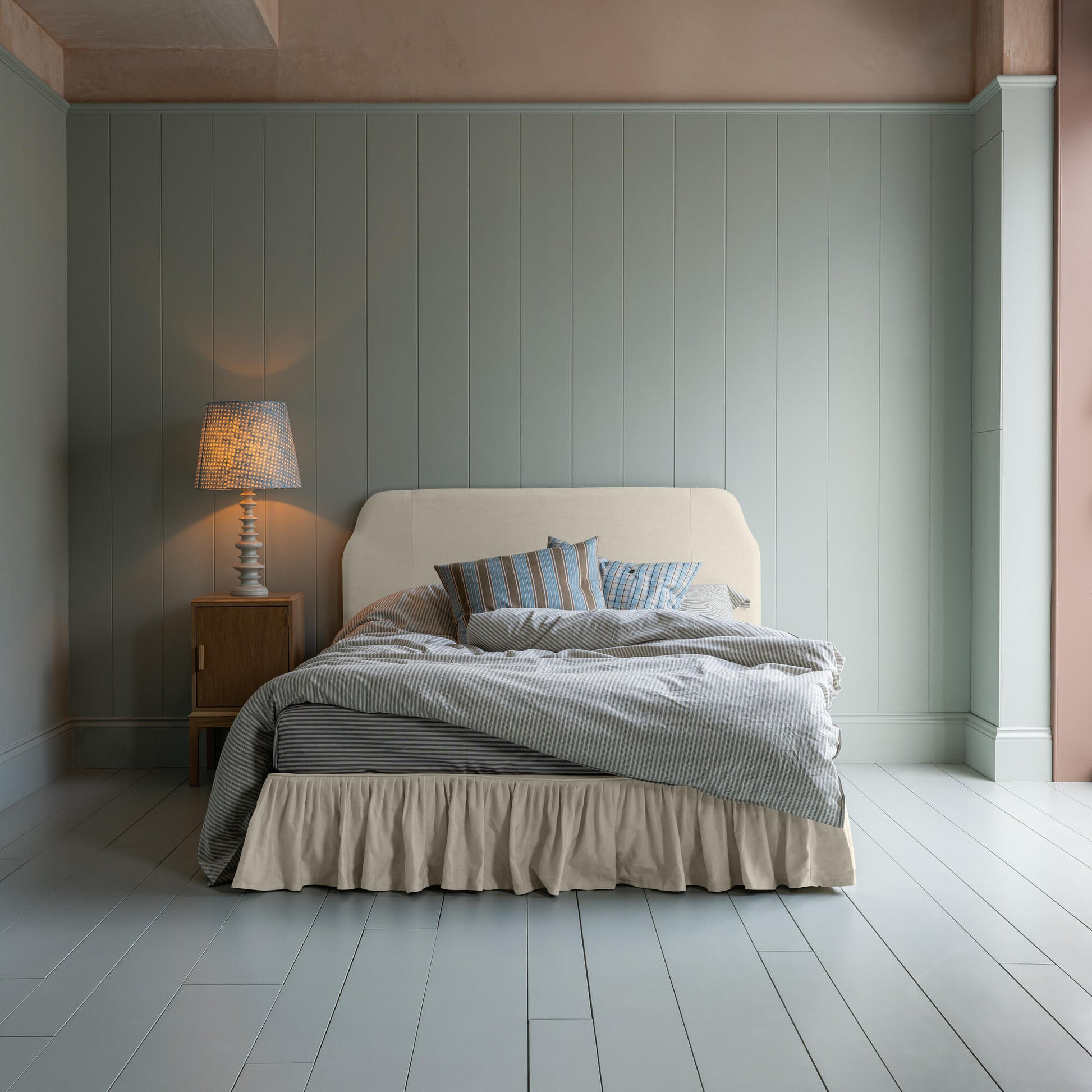 King-size upholstered bed in laidback linen dove with a ruffled valance, accompanied by a side table lamp, pillows, and a blanket.