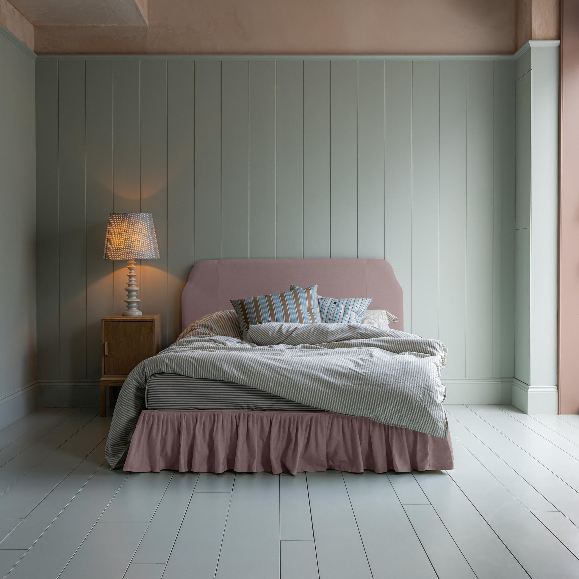 Upholstered bed in laidback linen heather with a ruffled valance, accompanied by a side table lamp, pillows, and a blanket.