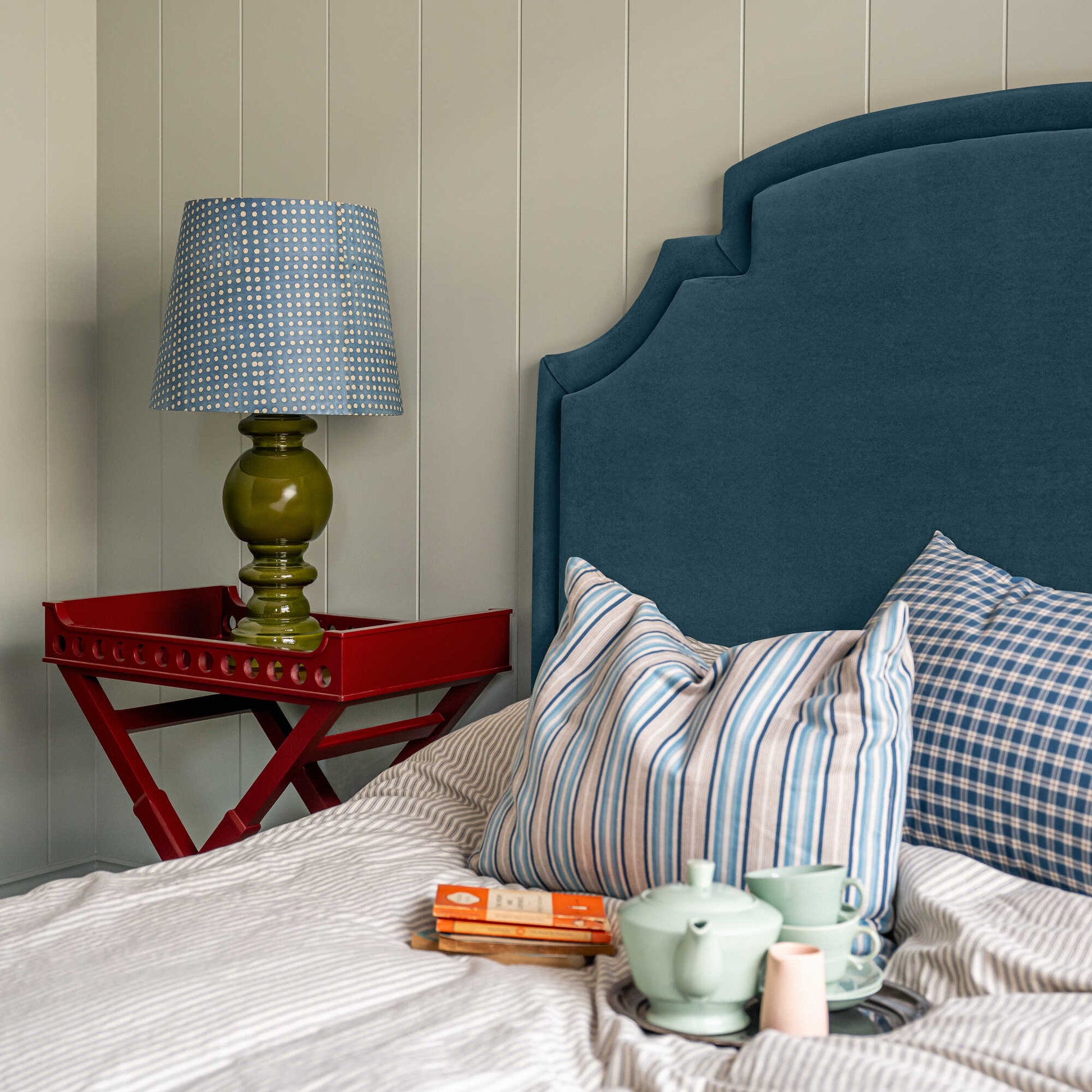 Seventh Heaven Upholstered Bed in Intelligent Velvet Aegean with Kick Pleat Valance, featuring a red side table with a lamp, books, and a teapot with cups on the bed, along with pillows.