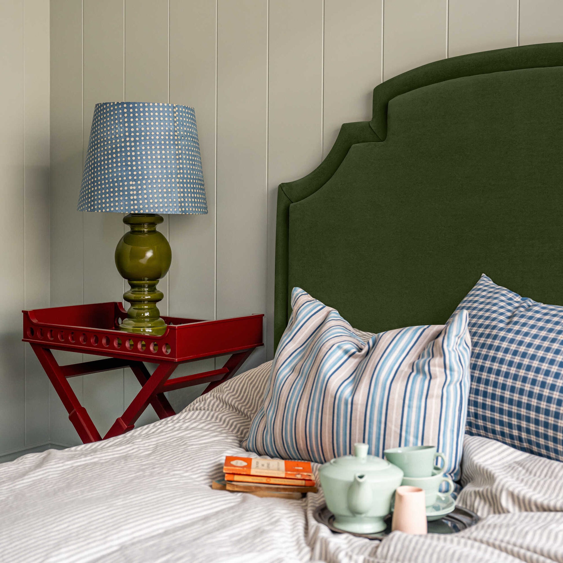 Seventh Heaven Upholstered Bed in Intelligent Velvet Juniper with Kick Pleat Valance, featuring a red side table with a lamp, books, and a teapot with cups on the bed, along with pillows.