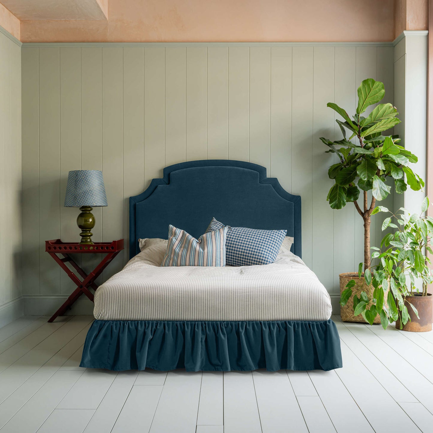 Seventh Heaven Upholstered Bed in Intelligent Velvet Aegean with Ruffled Valance, accompanied by a red side table with lamp, pillows, and a blanket.