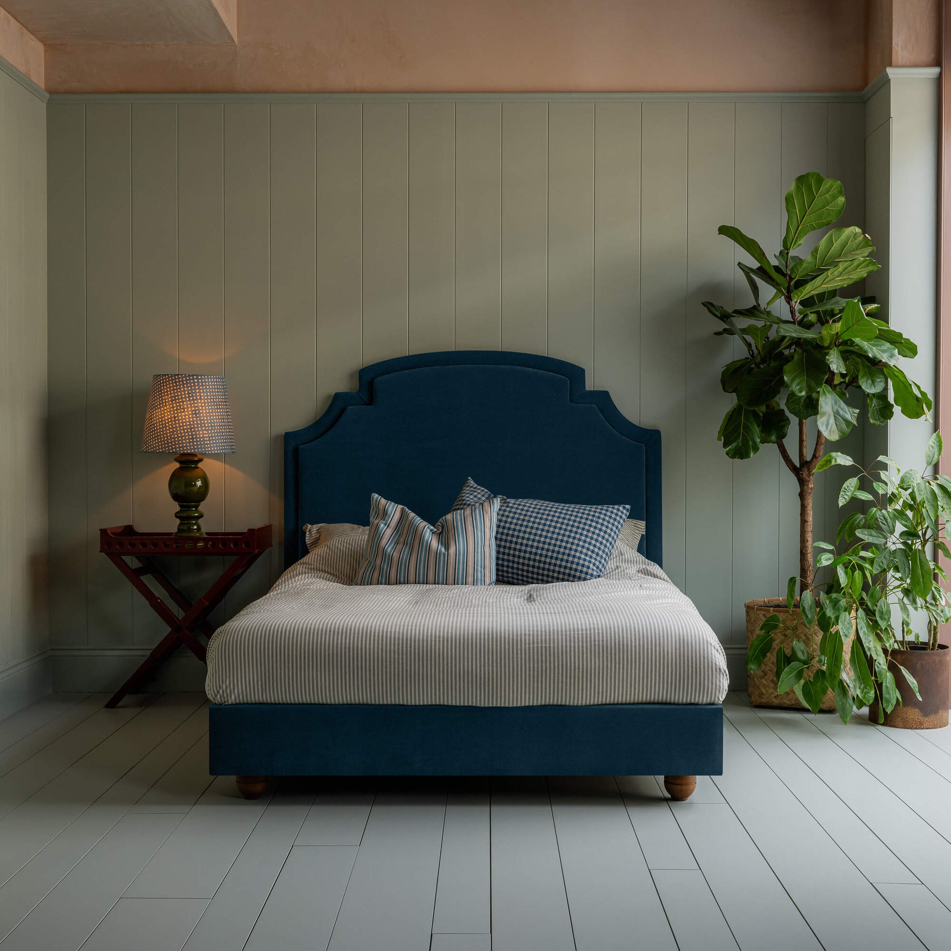 Upholstered bed in laidback linen heather with a ruffled valance, accompanied by a side table with lamp, indoor plant, pillows, and a blanket.