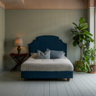  Upholstered bed in laidback linen heather with a ruffled valance, accompanied by a side table with lamp, indoor plant, pillows, and a blanket. 