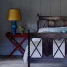  Ready Steady Tray Table in Berry Red with a Blue Base, paired with a Mustard Lampshade, designed by Nicola Harding & Co, placed beside the bed. 